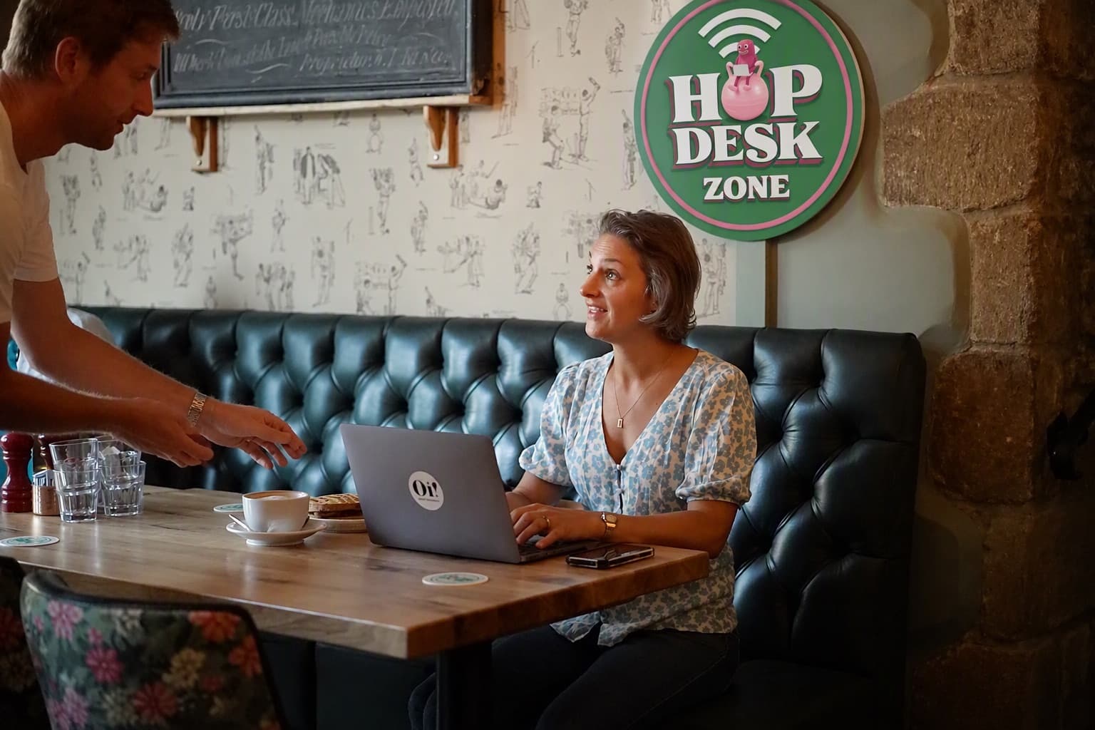 Someone working on a laptop at a pub table under a HopDesk Zone sign, as a coffee is set down beside them