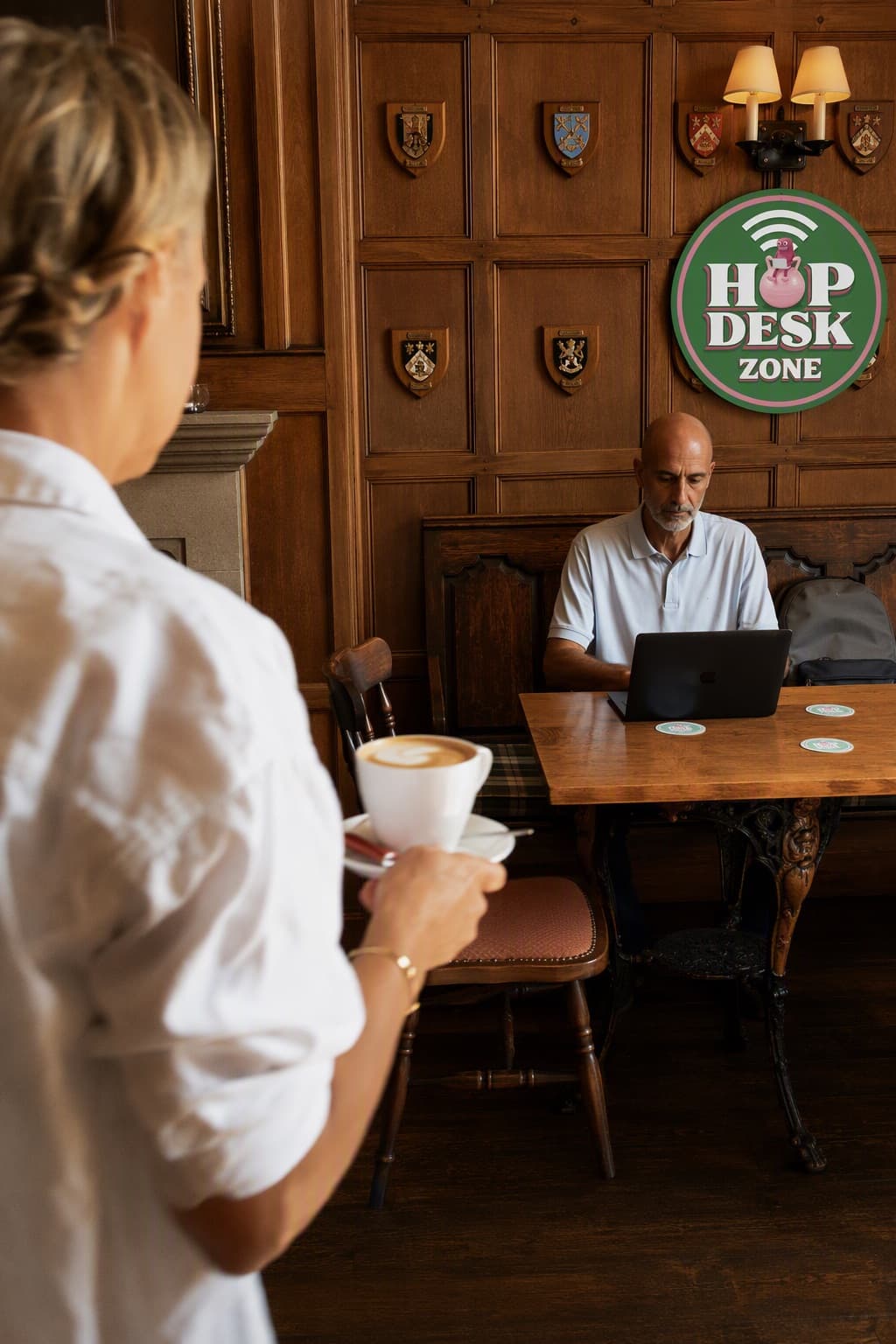 A coffee being placed next to an open laptop on a pub table, under a HopDesk Zone sign